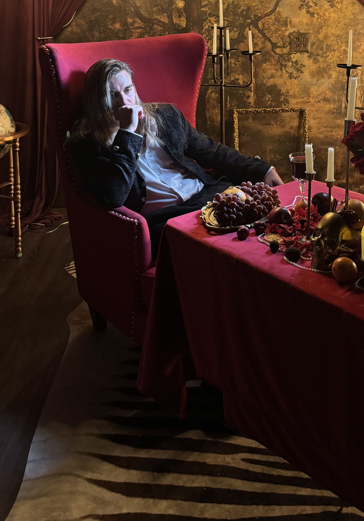 Jay Putty leaning back in a crimson chair beside a table of candles, fruit and roses