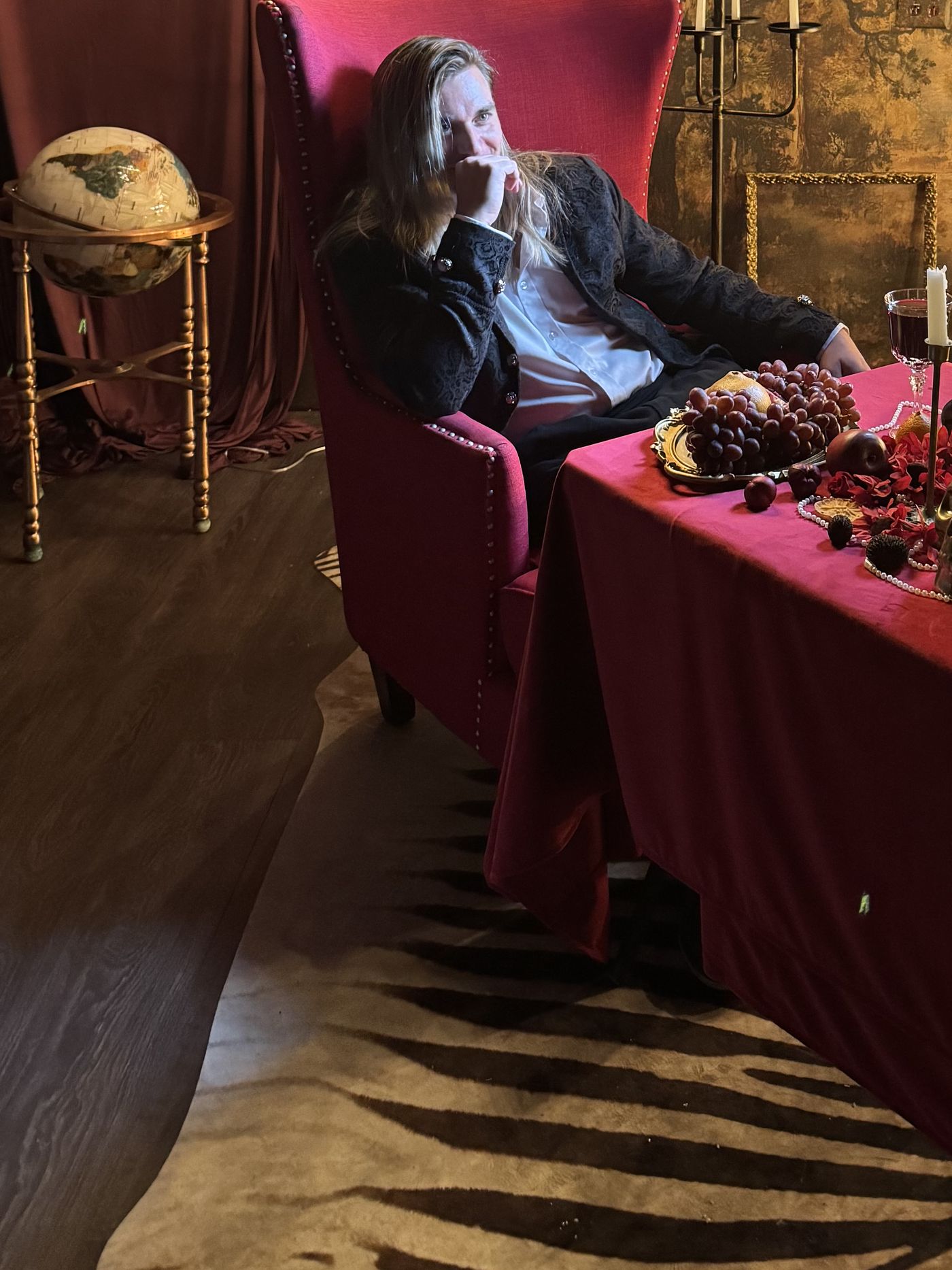 Jay Putty seated in a crimson chair, hand at his chin, candles and roses around him